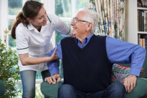 Two older adults sitting together in a bright, communal living room, one reading and one speaking with a caregiver.