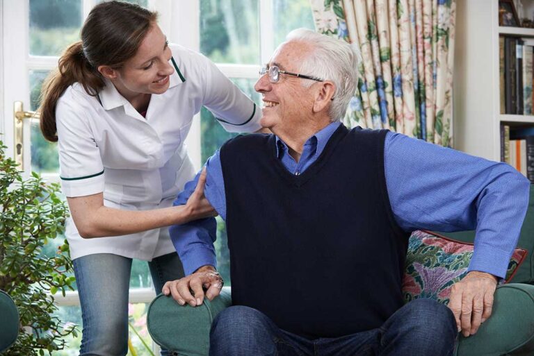 Two older adults sitting together in a bright, communal living room, one reading and one speaking with a caregiver.